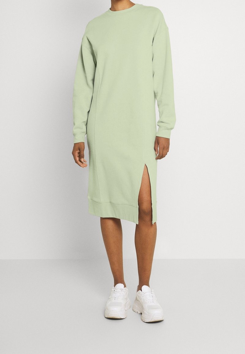 Personne portant une robe midi à manches longues vert clair avec une fente latérale, associée à des baskets blanches épaisses, sur un fond uni.