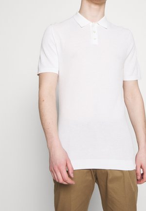 Mężczyzna ubrany w białą, teksturowaną koszulkę polo z krótkim rękawem i guzikami oraz beżowe spodnie, stojący na tle jednolitego jasnego tła.