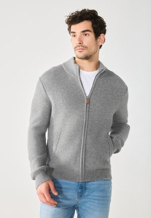 Grå strikket zip-up sweater med høj krave, med et tekstureret mønster, ribstrikkede ærmer og en lynlåstræk accent. Sammen med blå jeans.
