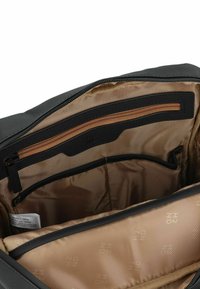 Interior de bolsa de tela negra con forro marrón claro, que cuenta con un bolsillo con cremallera y múltiples compartimentos para la organización. Incluye detalle del logotipo de la marca.