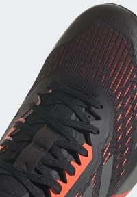 Close-up van een zwart en rode sportschoen met textiel, zwarte veters, en synthetische overlays op de neus en zijkanten.