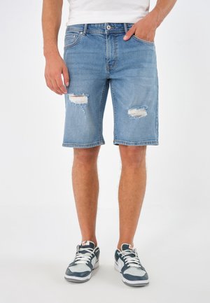 Mężczyzna w jasnoniebieskich, podartych szortach z denimu, białych i czarnych butach Nike, stojący z jedną ręką w kieszeni na białym tle.
