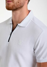 Hvid polo t-shirt med tekstureret stof, halv-lynlås foran og et subtilt geometrisk mønster. Den har en krave og et lille logo på brystet.