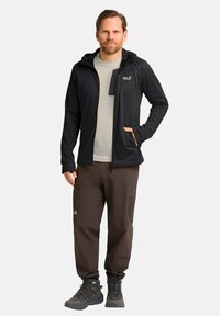 Giacca nera con zip e cappuccio, camicia grigia sotto e pantaloni marroni. Stivali di un grigio scuro con lacci texturizzati. Tasche notevoli sulla giacca e sui pantaloni.