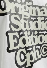 Valge kangast T-särk, millel on julge must ja kreemjas tekst retro fondis: "Original Studio Edition Cph@." Tekst on ülemõõtu ja erineva kõrgusega tähed.
