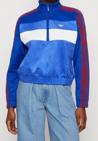 Blå och vit Adidas träningsjacka med röda ränder och logotyp på ärmarna, bärs med lösa ljusblå jeans.