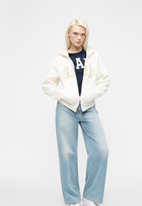 Νεαρή γυναίκα με ξανθά μαλλιά, που φοράει λευκό φούτερ GAP πάνω από μπλε σκούρο πουκάμισο, ανοιχτόχρωμα τζιν με φαρδιά μπατζάκια και λευκά αθλητικά παπούτσια, đứng με τα χέρια στις τσέπες.