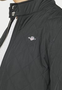 Black quilted jacket with snap collar, zipper closure, and small embroidered GANT logo on left chest over gray shirt.