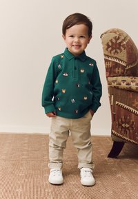 Žalias ilgomis rankovėmis polo marškinėlis su gyvūnų siuvinėjimu, smėlio spalvos kelnės ir balti sportbačiai. Paprastas dizainas su apykakle.