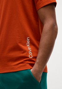 Oranžinė medvilninė marškinėliai su apvaliu iškirpimu ir trumpomis rankovėmis, kairėje pusėje esančiu baltu "Calvin Klein" logotipu.