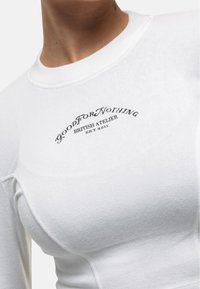Weißes Langarmoberteil aus strukturiertem Stoff mit einem gestickten schwarzen Logo, das auf der Brust "GOOD FOR NOTHING BRITISH ATELIER EST 2011" liest.