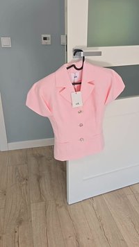 Rosa blus med korta, strukturerade ärmar, en spetsig krage och fem runda knappar. Har två framfickor och en hängande etikett.