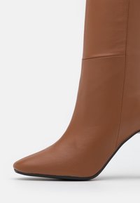 Bota de tacón alto de punta afilada en cuero marrón con textura suave y costuras visibles sobre un fondo blanco.