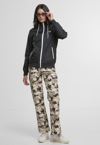 Black lightweight jacket with white accents, paired with beige camo cargo pants. Model wears reflective sunglasses and white sneakers.