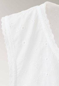 Blouse blanche sans manches avec broderie florale ajourée et bordure en dentelle aux manches et à l'encolure, présentée sur un mannequin.