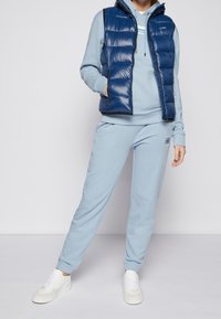 En person klädd i en ljusblå hoodie och matchande sweatpants med vita sneakers, samt en marinblå kviltad ärmlös väst mot en neutral bakgrund.