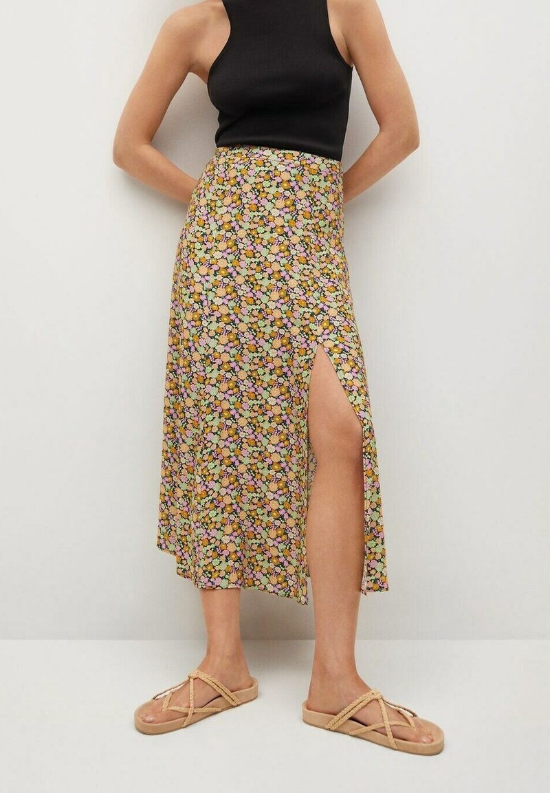Femme portant une jupe midi fleurie avec une fente haute sur le côté, un haut noir sans manches et des sandales à lanières en corde beige, debout contre un mur blanc.