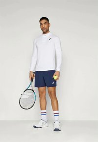Hvit langermet skjorte med krage, marineblå shorts, hvite joggesko med blå detaljer, holder en tennisracket og ball.