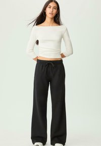 Weißes Langarmshirt mit einem Bootsausschnitt, kombiniert mit schwarzen, weit geschnittenen Sweatpants. Das Outfit hat eine Kordelzugtaille und Taschen.