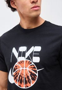 Νέος άνδρας που φοράει μαύρο T-shirt Nike με λευκό λογότυπο και πορτοκαλί-μαύρο γραφικό μπάσκετ στο μπροστινό μέρος.