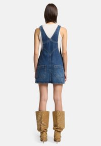 Abito in denim con un corpetto aderente, spalline larghe e due grandi tasche frontali. Indossato sopra una maglietta bianca, abbinato a stivali perforati al ginocchio.
