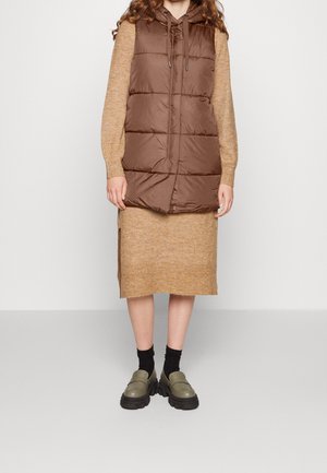 Persoon draagt een bruin gewatteerd vest over een zandkleurige gebreide jurk, zwarte sokken en olijfgroene chunky loafers, staand tegen een effen achtergrond.