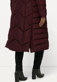Parka bordeaux con design a zig zag, con fessura laterale e texture liscia. Abbinato a pantaloni neri aderenti e stivali con tacco.