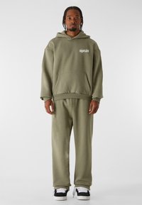 Hoodie vert olive avec poche avant et logo, associé à un jogger assorti. Les deux sont en tissu doux et texturé, avec des baskets blanches.