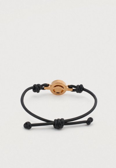 Juodas virvelės apyrankė su blizgančiu aukso atspalvio metaliniu užsegimu. Užsegimas yra apvalus, lygios tekstūros, o virvelėje yra mazgų akcentai.