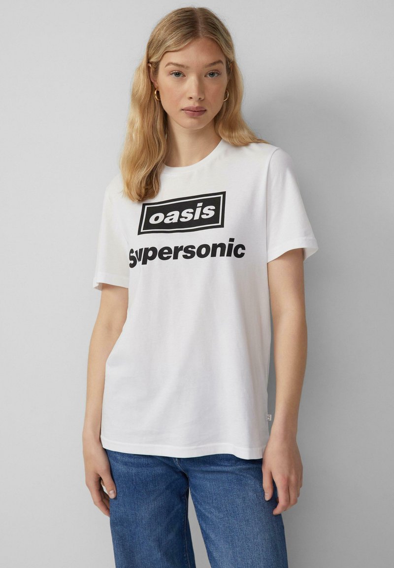 Weißes Baumwoll-T-Shirt mit einem schwarzen Grafikdruck, der die Worte "oasis" und "supersonic" zeigt, kombiniert mit blauen Jeans.