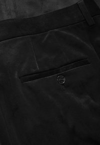 Gros plan sur un pantalon en daim noir avec une poche passepoilée à l'arrière fermée par un seul bouton noir et une attache de ceinture au-dessus.