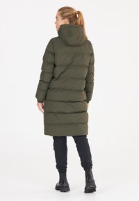 Abrigo acolchado verde oliva con capucha, que presenta secciones acolchadas horizontales. Lleva puesto sobre pantalones negros ajustados y botas negras hasta el tobillo.