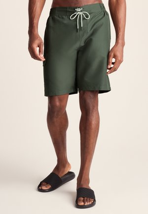 Man wearing olive green swim trunks with drawstring and black slide sandals, standing against a plain light background, visible from waist down.