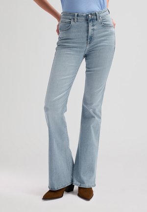 Osoba ubrana w jasnoniebieskie, wysokie dzwony jeansowe i brązowe botki na obcasie z zakończeniem w szpic na neutralnym tle.