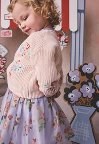 Toddler with curly hair wearing a pink sweater with embroidered flowers and a floral skirt, smiling sideways against a floral backdrop.