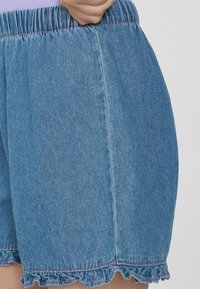 Džínové šortky s elastickým pasem, volánkovým lemem a růžovým prošitím. Materiál je světle modrý a má texturovaný povrch.