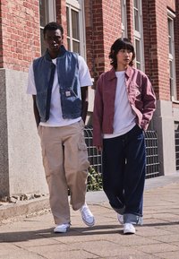 Two models walk side by side. One wears a denim vest over a white t-shirt and light beige cargo pants. The other wears a pink jacket, white t-shirt, and dark jeans. Both have white sneakers.