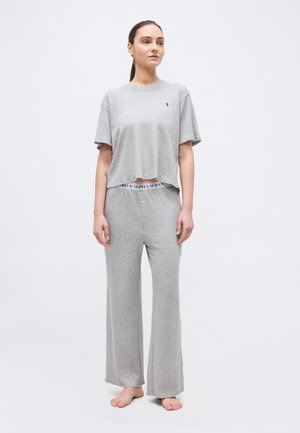 Vrouw staat op blote voeten en draagt een lichtgrijs cropped T-shirt en bijpassende losse loungebroek tegen een effen achtergrond.