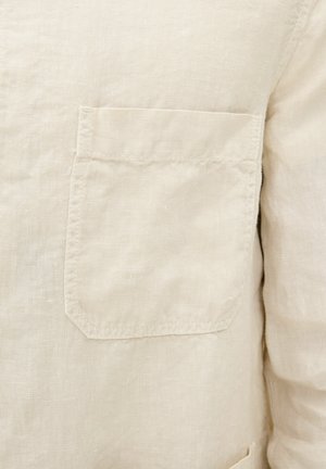 Beige linnen shirt met een patch pocket, met een gestructureerde stof en zichtbare stikdetails. Het ontwerp is eenvoudig en functioneel.