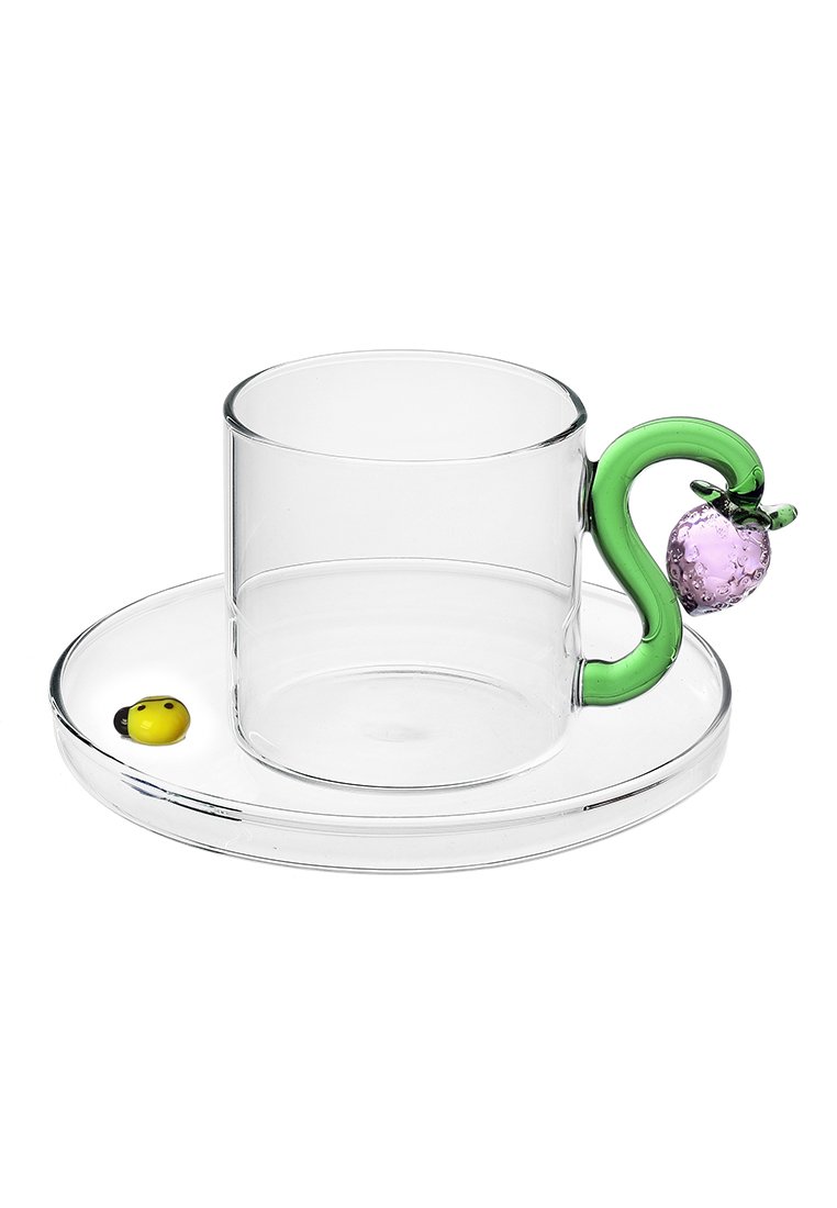 Przezroczysty szklany kubek z zielonym, zakrzywionym uchwytem z fioletowym akcentem i żółtą biedronką na okrągłej, przezroczystej podstawce.