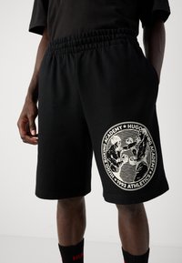 Svart shorts med elastisk midje, med en hvit sirkulær "Hugo Boxing Academy 1993 Athletics" logo med to boksere i aksjon.