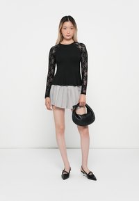 Haut noir à manches longues avec des détails en dentelle, taille peplum, associé à une jupe grise plissée et un sac à main noir. Silhouette mince et moderne.