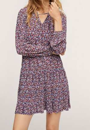 Kobieta ubrana w bordo sukienkę z długim rękawem, sięgającą kolan, z niebiesko-białym kwiatowym wzorem i małym wycięciem w kształcie litery V przy dekolcie.