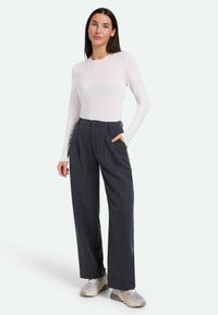 Weißes Langarmoberteil kombiniert mit dunkelgrauen Nadelstreifen-Hose mit weitem Bein, die über Seitentaschen und einen Knopfverschluss verfügt. Graue Sneaker.