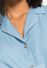 Světle modrá denimová košile s límcem a zapínáním na knoflíky. Tkanina má jemnou strukturu s mírnými variacemi odstínu.