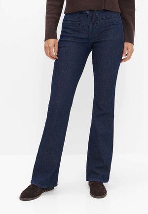 Person iført mørkeblå bootcut jeans, brune sko og en brun langærmet top, stående mod en hvid baggrund.