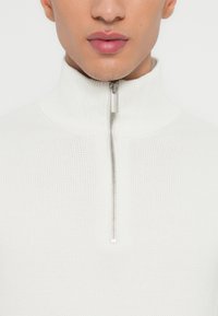 Mann in einem weißen, gerippten Strickpullover mit hohem Kragen und halbem Reißverschluss, der das untere Gesicht und den Hals zeigt.