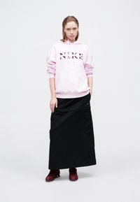 Rozā kapuces džemperis ar 'NIKE' logo, papildināts ar melnu garu svārku ar kabatām. Modelis valkā bordo kedas.