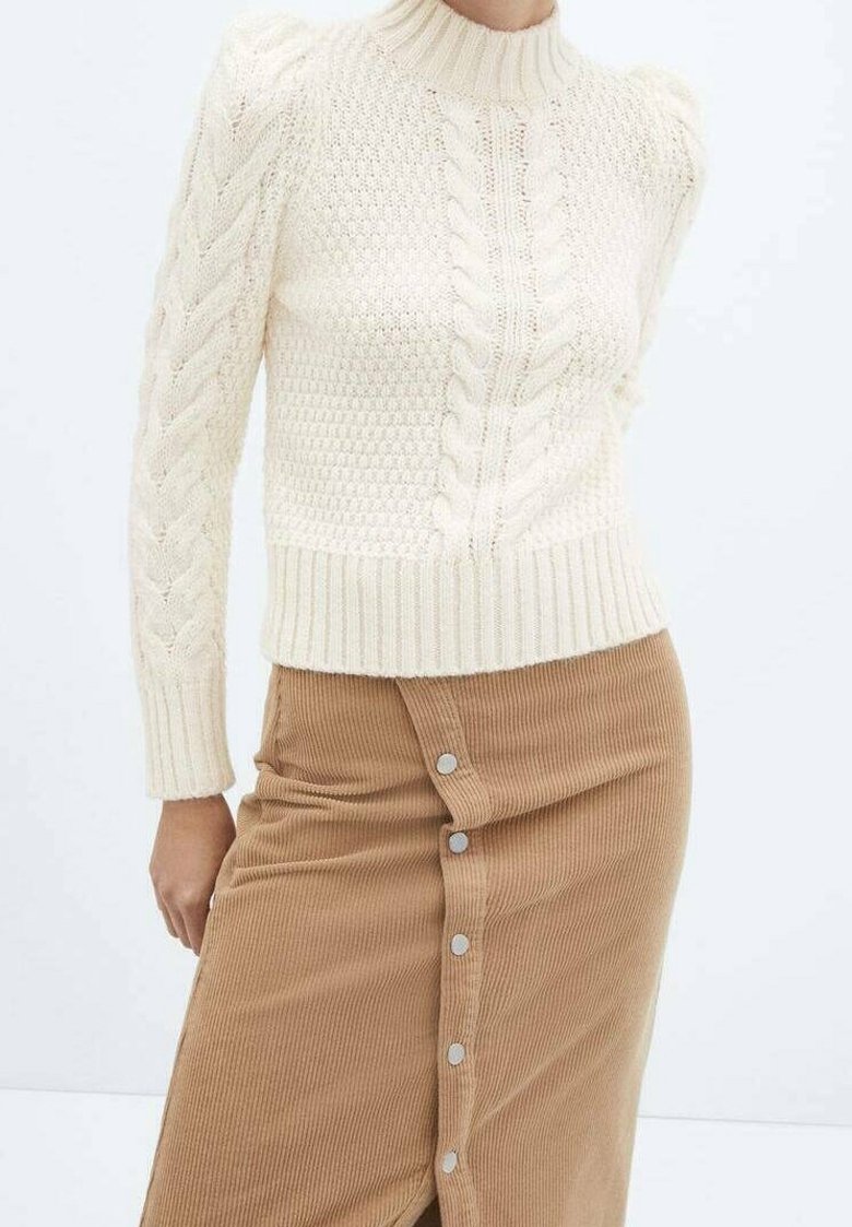 Woman wearing cream cable-knit turtleneck sweater and high-waisted tan corduroy skirt with front silver buttons, standing against white background.