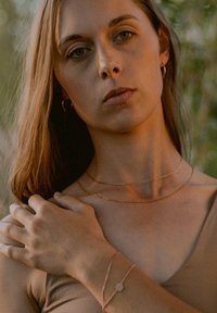 Layered rose gold necklaces and a bracelet with a circular charm; model has long brown hair and a beige top. Natural greenery in background.